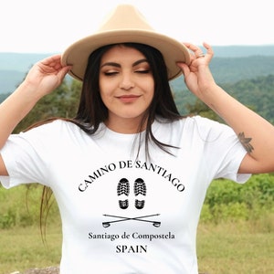 May include: White t-shirt with black text that reads "Camino de Santiago" with a graphic of two footprints and two walking sticks. Below the graphic is the text "Santiago de Compostela SPAIN".