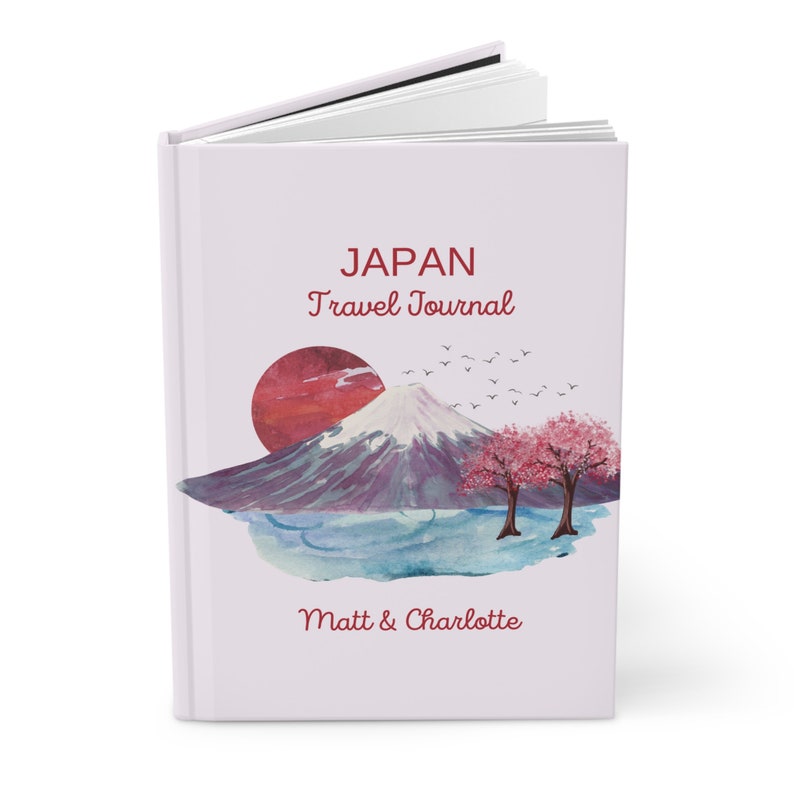 Op de afbeelding: Een wit reisdagboek met een aquarel-illustratie van de Mount Fuji, een rode zon, kersenbloesems en vogels. De tekst "JAPAN Travel Journal" en "Matt & Charlotte" staat op de omslag gedrukt.