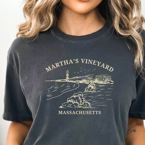 May include: A dark gray t-shirt with a white graphic design of a lighthouse, beach, and sailboats. The text "Martha's Vineyard Massachusetts" is printed on the shirt.