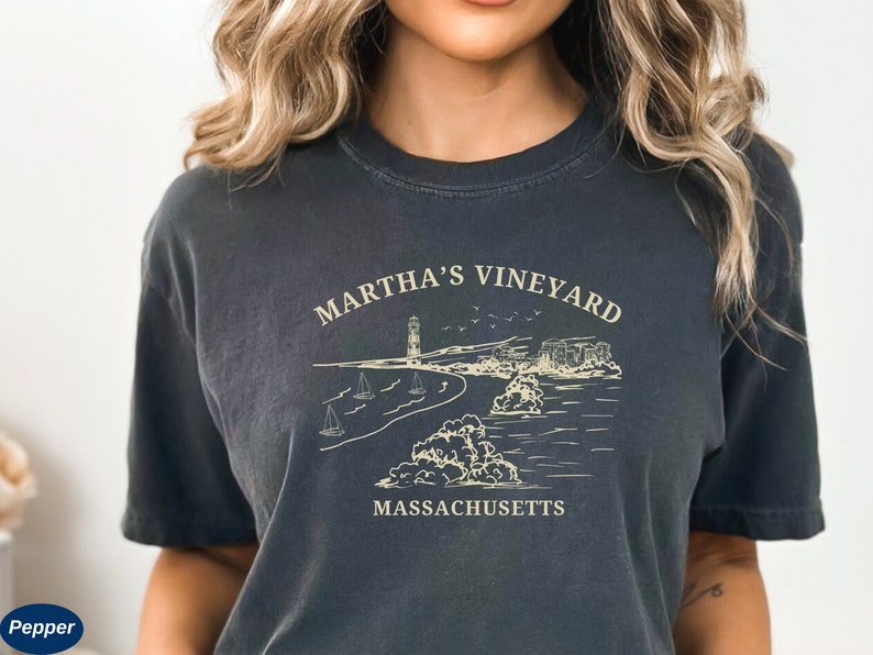 May include: A dark gray t-shirt with a white graphic design of a lighthouse, beach, and sailboats. The text "Martha's Vineyard Massachusetts" is printed on the shirt.