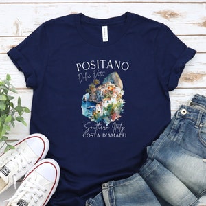 May include: Navy blue t-shirt with a white graphic design featuring a watercolor illustration of Positano, Italy. The text "Positano Dolce Vita Southern Italy Costa D'Amalfi" is printed on the shirt.