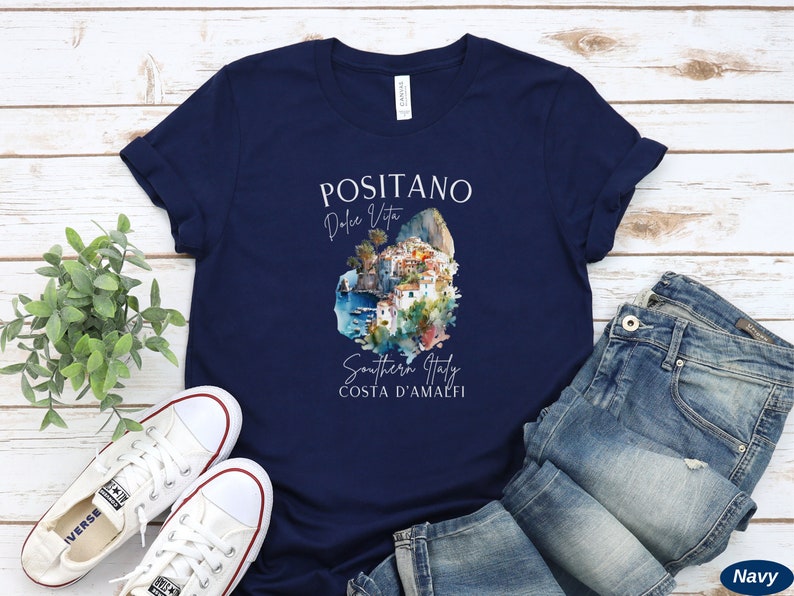 May include: Navy blue t-shirt with a white graphic design featuring a watercolor illustration of Positano, Italy. The text "Positano Dolce Vita Southern Italy Costa D'Amalfi" is printed on the shirt.