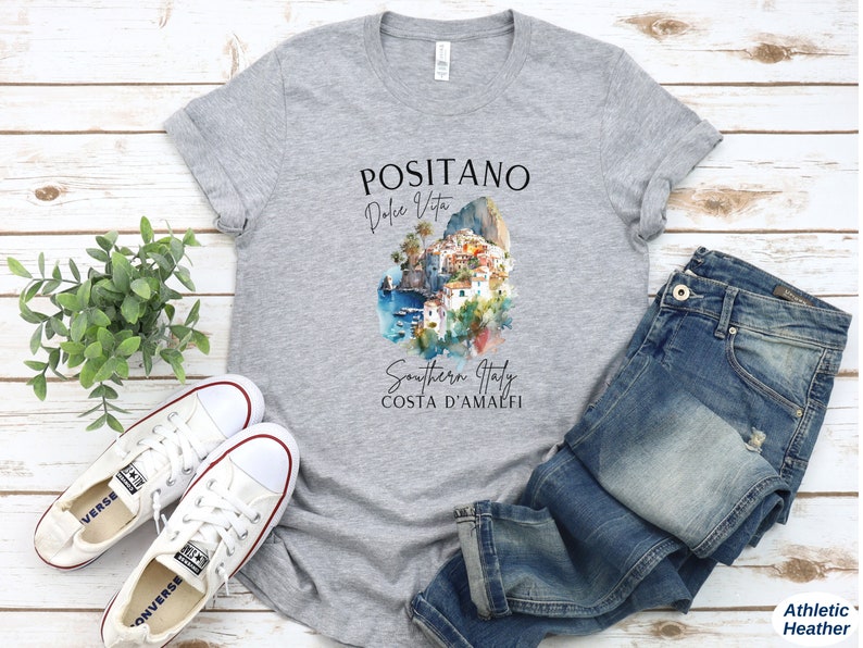 May include: A gray t-shirt with a watercolor illustration of Positano, Italy. The text on the shirt reads "Positano Dolce Vita Southern Italy Costa d'Amalfi".