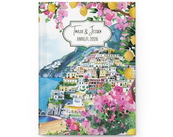 Op de afbeelding: Een hardcover-dagboek met een aquarelillustratie van Amalfi, Italië. De omslag toont een kustplaats met kleurrijke gebouwen, roze bloemen, citroenen en de tekst "Taylor & Jessica, Amalfi, 2026" in een decoratief kader.