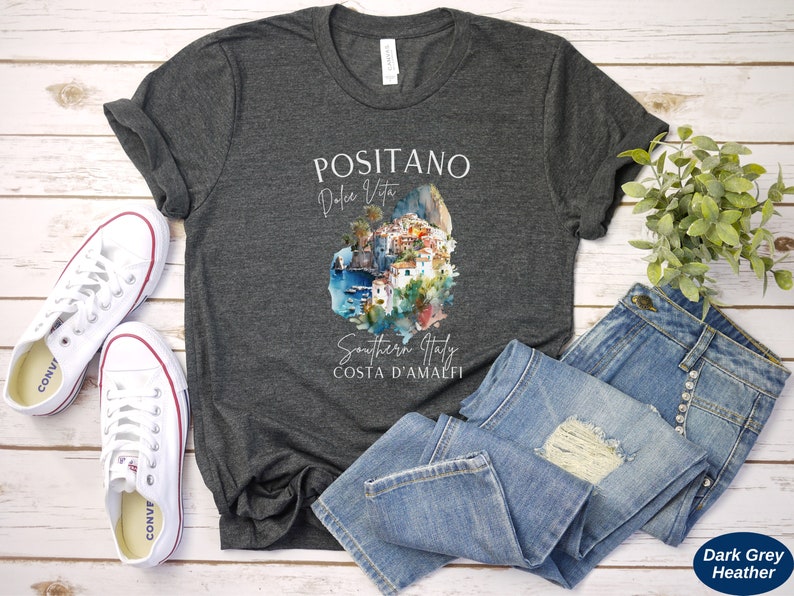 May include: Dark grey heather t-shirt with a watercolor illustration of Positano, Italy. The text "Positano, Dolce Vita, Southern Italy, Costa d'Amalfi" is printed on the shirt.