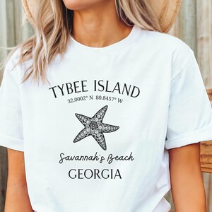 May include: White t-shirt with black text that reads "Tybee Island 32.0002&deg; N 80.8457&deg; W Savannah's Beach GEORGIA" and a black and white starfish graphic.