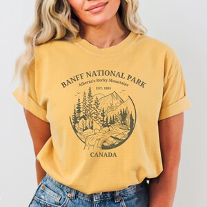 May include: A mustard yellow t-shirt with a graphic print of a mountain landscape and the text "BANFF NATIONAL PARK Alberta's Rocky Mountains EST. 1885 CANADA".