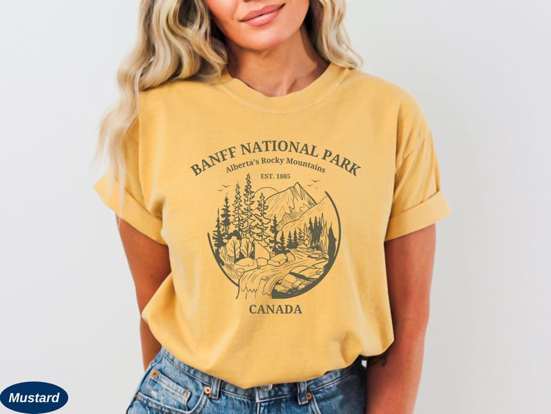 May include: A mustard yellow t-shirt with a graphic print of a mountain landscape and the text "BANFF NATIONAL PARK Alberta's Rocky Mountains EST. 1885 CANADA".