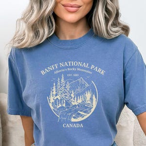 May include: A light blue t-shirt with a graphic print of a mountain scene and the text "Banff National Park, Alberta's Rocky Mountains, Est. 1885, Canada".