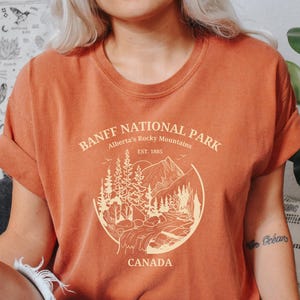 May include: A rust-colored t-shirt with a graphic design of a mountain landscape and the text "Banff National Park, Alberta's Rocky Mountains, Est. 1885, Canada".
