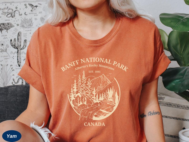 May include: A rust-colored t-shirt with a graphic design of a mountain landscape and the text "Banff National Park, Alberta's Rocky Mountains, Est. 1885, Canada".