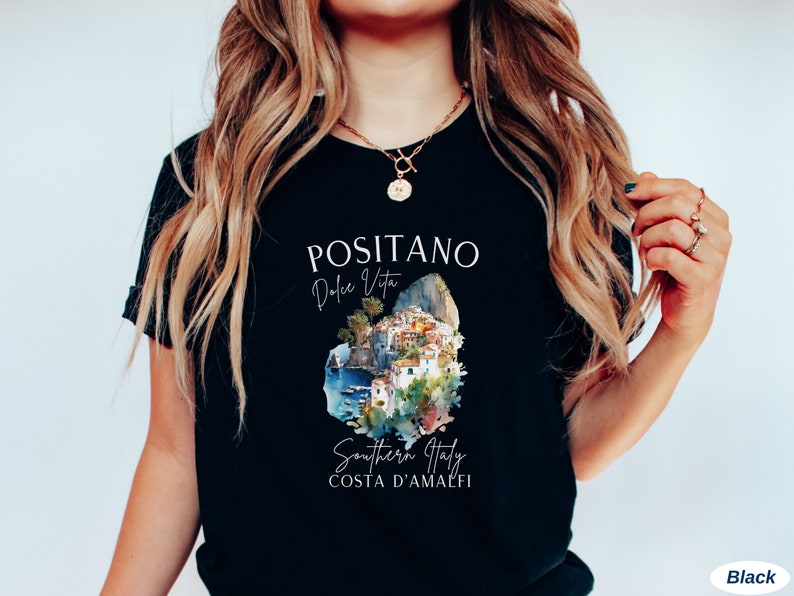 May include: Black t-shirt with a white graphic design featuring the text "Positano Dolce Vita" and a watercolor illustration of the town of Positano, Italy. The text "Southern Italy Costa d'Amalfi" is also included in the design.