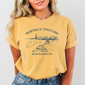 May include: A mustard yellow t-shirt with a graphic of a lighthouse, beach, and sailboats. The text "Martha's Vineyard, Massachusetts" is printed on the shirt.
