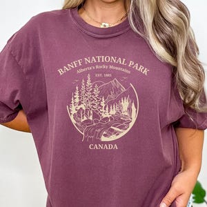 May include: A maroon t-shirt with a graphic design of a mountain scene and the text "Banff National Park, Alberta's Rocky Mountains, Est. 1885, Canada".