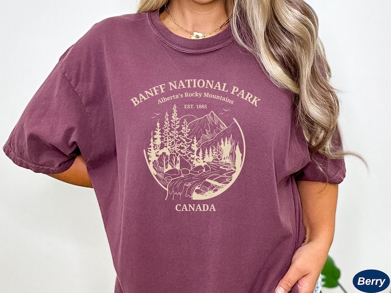 May include: A maroon t-shirt with a graphic design of a mountain scene and the text "Banff National Park, Alberta's Rocky Mountains, Est. 1885, Canada".