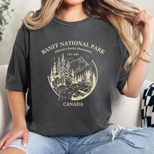 May include: A dark gray t-shirt with a vintage-style graphic of Banff National Park, Alberta's Rocky Mountains. The graphic includes a mountain range, trees, and a river. The text "Banff National Park", "Alberta's Rocky Mountains", "Est. 1885", and "Canada" is printed in white.