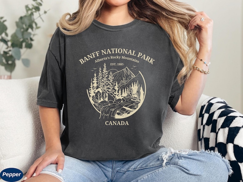 May include: A dark gray t-shirt with a vintage-style graphic of Banff National Park, Alberta's Rocky Mountains. The graphic includes a mountain range, trees, and a river. The text "Banff National Park", "Alberta's Rocky Mountains", "Est. 1885", and "Canada" is printed in white.