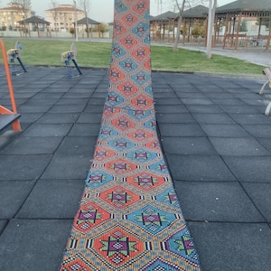 May include: A long, narrow rug with a colorful geometric pattern in shades of orange, blue, green, and purple. The rug has a fringe on both ends.