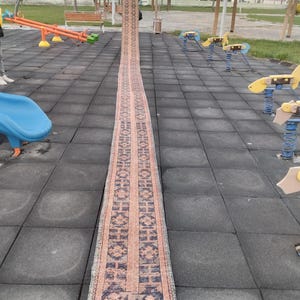 Puede incluir: Una alfombra larga y estrecha con un patrón geométrico rosa y azul. La alfombra está colocada sobre una superficie de caucho negro en un patio de recreo.