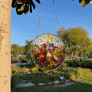 May include: A stained glass suncatcher featuring a colorful scene of three chickens in a field with a church in the background. The suncatcher is round and has a chain for hanging.