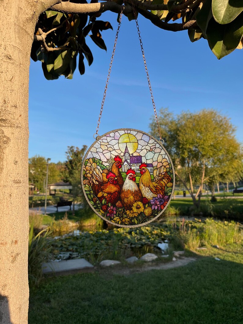 May include: A stained glass suncatcher featuring a colorful scene of three chickens in a field with a church in the background. The suncatcher is round and has a chain for hanging.