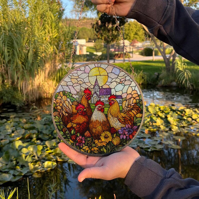 May include: A stained glass window hanging features a colorful scene of three chickens in a field of flowers with a church in the background.