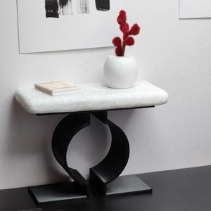 May include: A white marble console table with a black metal base. The table has a unique design with a circular opening in the base. A small book and a white vase with a red plant are on the table.