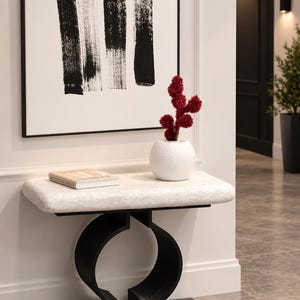 May include: A console table with a rectangular white textured top and a black base featuring two curved, omega-shaped supports. A white spherical vase containing a dark red fuzzy plant sits on the table next to a book with a light-colored cover.