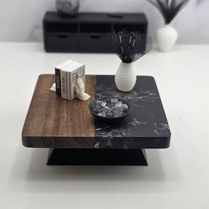 May include: A square miniature coffee table featuring a split design with one half in dark brown wood grain and the other in black marble pattern. It holds three small books, a white vase with black decorative stems, and a black bowl of crystals.