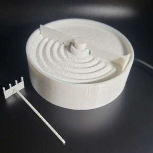 May include: White tabletop zen garden with a circular base and a small rake. The base has a rotating section with white sand and a small white plastic tool to create patterns in the sand.