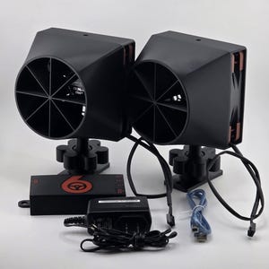 May include: Two black angled fan units with copper-coloured accents, mounted on black bases. A black control box with a red logo, a power adapter, and cables are also present. The fans have a circular design with multiple blades.