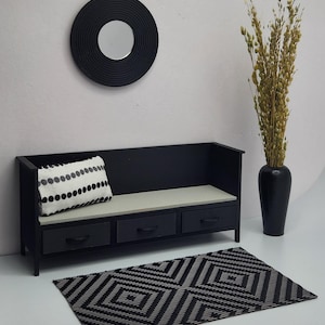 May include: Black bench with three drawers and a white and black patterned pillow. The bench is in front of a black and white patterned rug and a black round mirror with a white mirror inside. A vase with dried flowers is on the right side of the image.