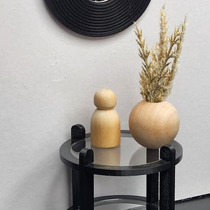 May include: A black round mirror with a concentric circle design sits above a black two-tiered side table. A wooden doll and a wooden vase with dried flowers are on the table.