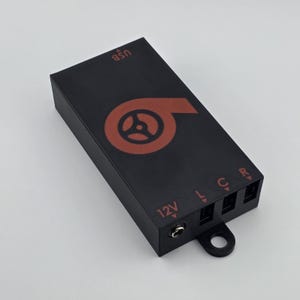May include: Black rectangular electronic device with a red logo and text. The logo is a stylized swirl with a steering wheel inside. Text includes "USB", "12V", "L", "C", and "R". A small loop is attached to the bottom.