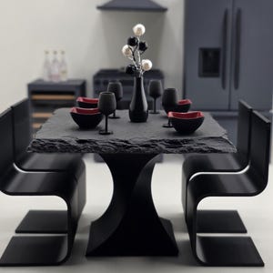 May include: A miniature dining set featuring a square, rough-edged black slate tabletop supported by a curved pedestal base. Four black chairs accompany the table, which is set with black and red bowls, black wine glasses, and a central vase.