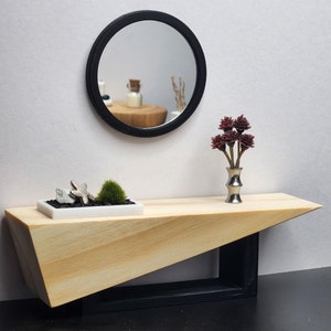 May include: A wooden table with a black metal frame and a triangular top. The table has a black round mirror with a reflective surface hanging above it. A small white planter with a starfish and green plants sits on the table. A vase with red flowers sits on the table.