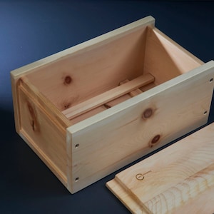 Könnte beinhalten: Eine hellfarbene Holzkiste mit abnehmbarem Deckel. Die Kiste ist rechteckig mit einer natürlichen Holzmaserung. Der Deckel ist teilweise geöffnet und gibt den Blick ins Innere frei. Die Kiste wird wahrscheinlich zur Aufbewahrung oder Präsentation verwendet.