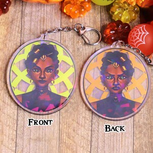 May include: A round, clear acrylic keychain with a colorful image of a woman with dark skin, dark hair, and gold jewelry. The front of the keychain features a yellow background with a geometric pattern. The back of the keychain features an orange background with a geometric pattern. The keychain is labeled "FRONT" and "BACK".