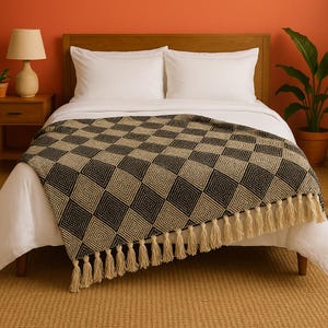 May include: A black and tan geometric patterned throw blanket with a fringe of tassels. The blanket is draped over a bed with white sheets and pillows. The headboard is wooden, and the background is an orange wall.