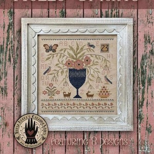 May include: A framed cross-stitch sampler with a floral design, a butterfly, and birds. The words "HELLO SPRING" are at the top. The frame is white with a scalloped edge. The sampler includes the text "featuring 8 designs".