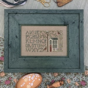 Könnte beinhalten: Ein gerahmter Kreuzstich-Sampler mit einem türkisfarbenen Rahmen. Der Sampler zeigt ein Haus mit einer roten Tür, einen Baum und das Alphabet in einer Vintage-Schrift. Der Sampler befindet sich auf einem floralen Stoffhintergrund.