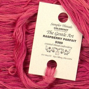 Puede incluir: Una bobina de hilo de bordar de color rosa con la etiqueta "Sampler Threads COLORFAST The Gentle Art RASPBERRY PARFAIT 0380 counted thread embroidery PO Box 670 New Albany, Ohio 43054"