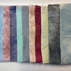 Puede incluir: Una colección de diez piezas de tela rectangulares en varios colores, incluyendo amarillo, azul, rosa, morado, turquesa, gris, beige, burdeos, verde claro y gris oscuro. Cada pieza está doblada y tiene una apariencia texturizada, con costuras visibles en el borde superior.