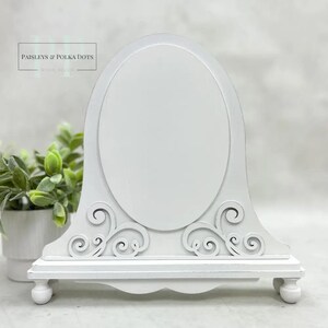 May include: White decorative clock-shaped frame with an oval center and ornate scrollwork. The frame has a shelf at the bottom and rests on four small, rounded feet. The text "Paisleys & Polka Dots" is visible in the upper left corner.