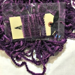 May include: A close-up shot of a bundle of purple trim with a pom-pom edge. The trim is packaged in a clear plastic bag with two cream-colored tags and a black ribbon. The trim is a deep purple color with a lighter purple accent.