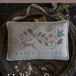 May include: A cross stitch sampler pattern featuring a bird, flowers, and the text "Hello Spring". The pattern is on a beige cushion with a white beaded border. The cushion is sitting on a wooden tray with buttons and scissors.