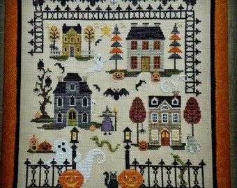 May include: Cross-stitch pattern featuring a spooky Halloween village with houses, trees, bats, ghosts, pumpkins, and a witch. The pattern is framed with a black fence and a black border.