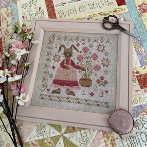 Könnte beinhalten: Ein gerahmtes Kreuzstich-Design mit einem Hasen, der eine Gießkanne und einen Blumentopf hält. Das Design ist von einem Rand aus Blumen und Sternen umgeben. Der Text "Stitch by Stitch" ist in der oberen rechten Ecke des Bildes sichtbar.