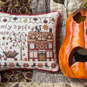 Puede incluir: Una exhibición de Halloween con un cojín de punto de cruz con el texto "Candy Apple House", una calabaza naranja tallada y dos calabazas de cerámica. El cojín tiene un borde rojo y un diseño detallado de la casa.