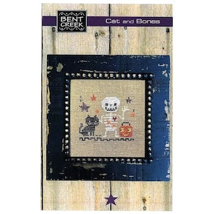 May include: A cross-stitch pattern featuring a skeleton holding a jack-o-lantern and a black cat. The pattern is framed in a black wood frame with a gold trim. The title "Cat and Bones" is printed above the frame.